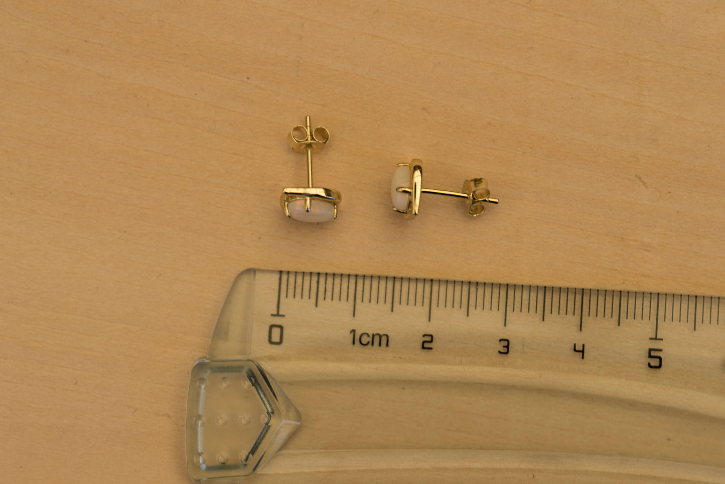 Gold stud opal earrings with a ruler for scale on a wooden surface