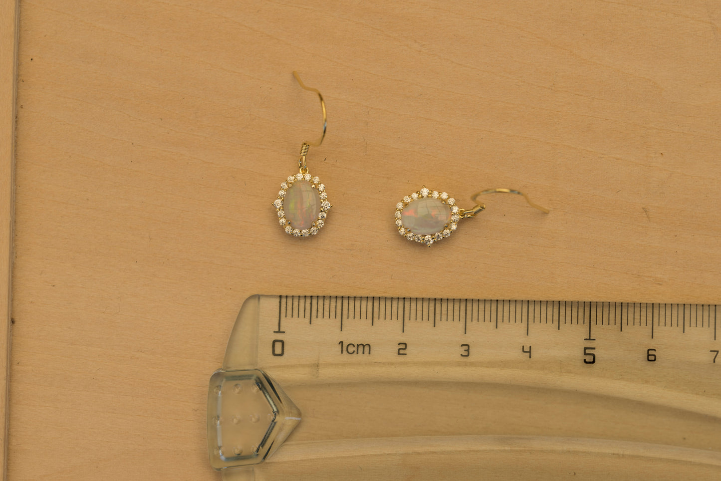 Pair of gold earrings with opal stones on a beige background with a ruler for scale.
