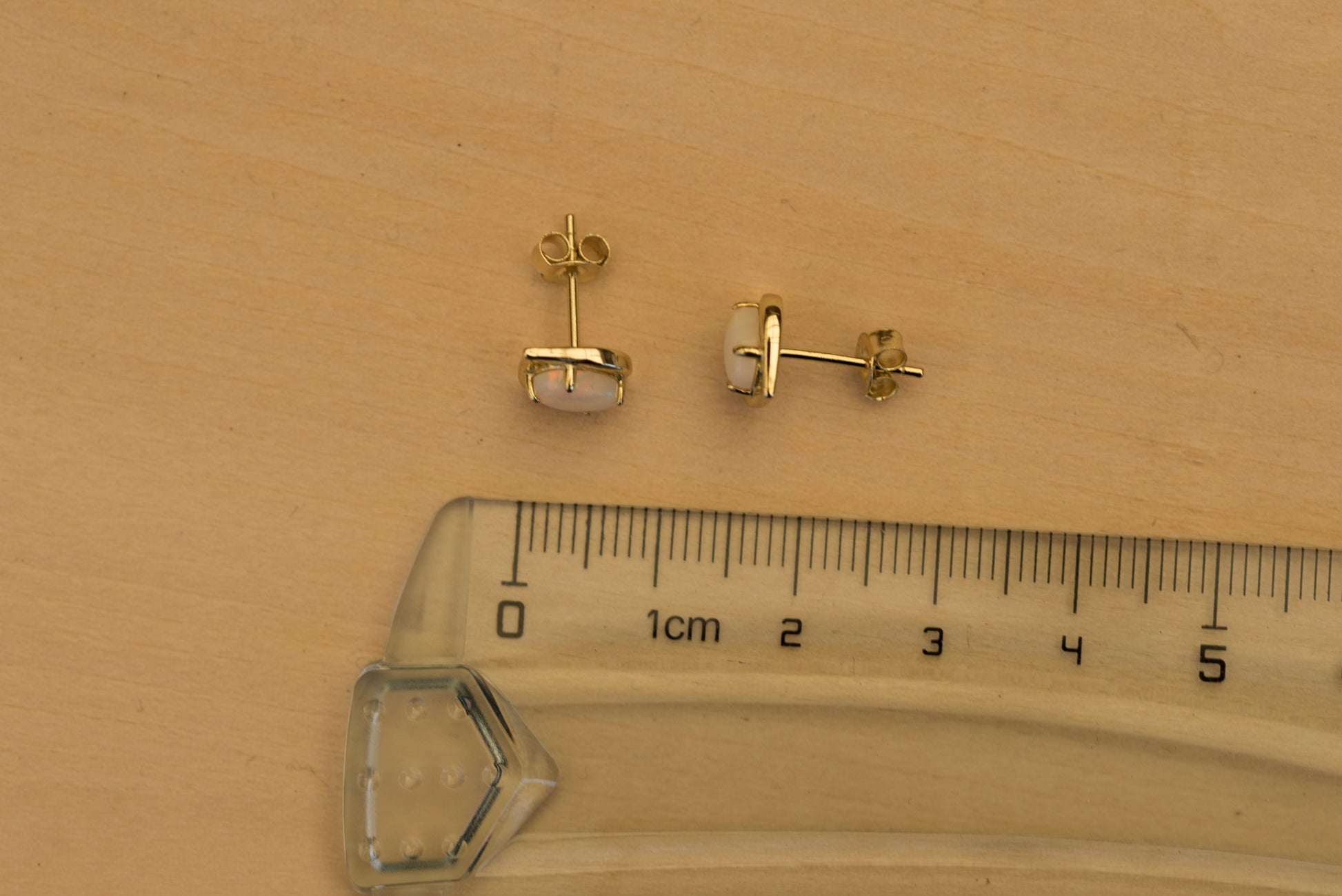Gold stud opal earrings with a ruler for scale on a wooden surface