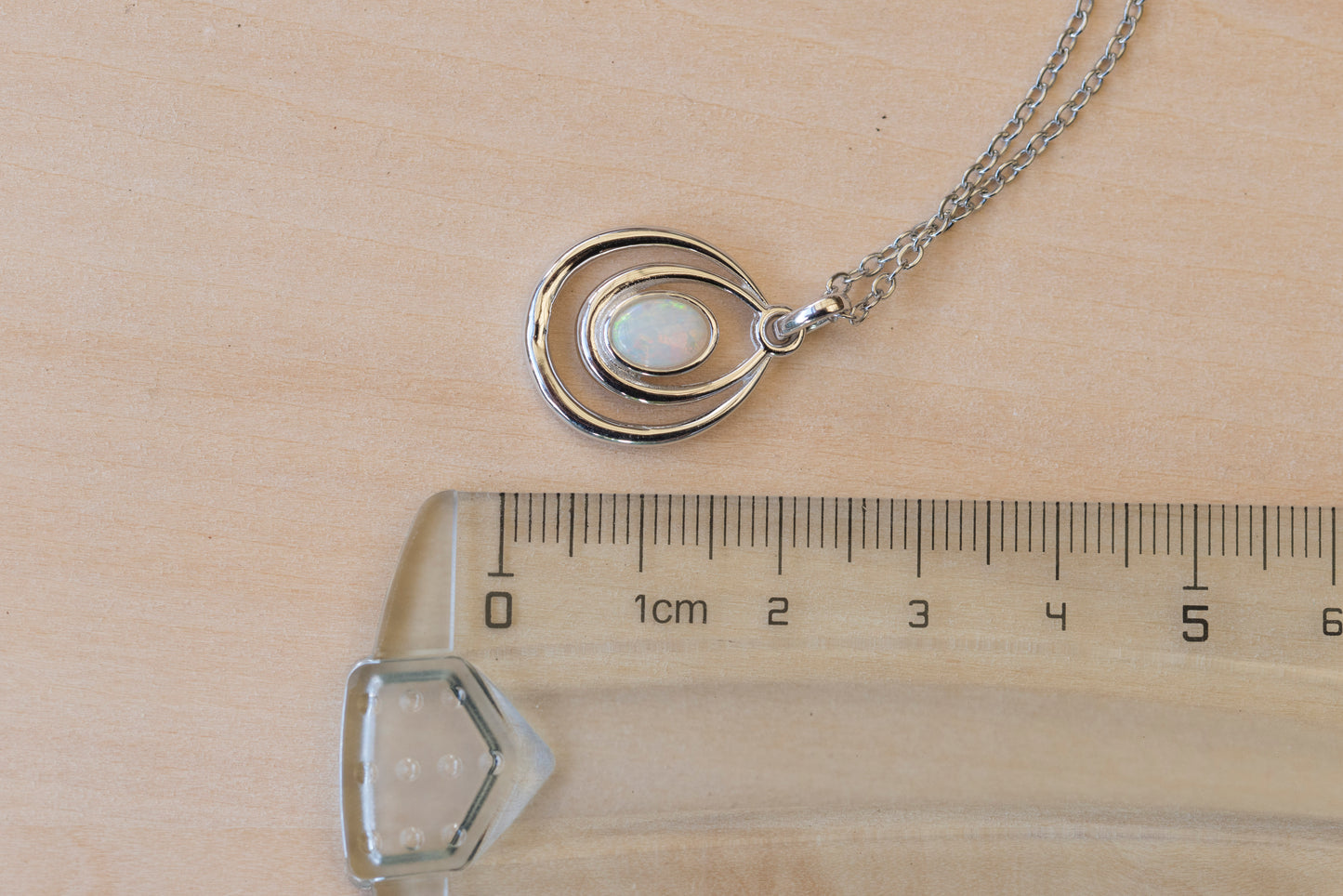 Silver necklace with a pendant on a wooden surface next to a ruler for scale.